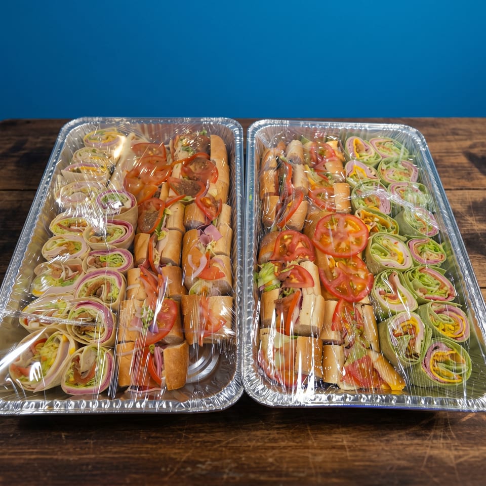 Two catering trays of assorted subs and wraps, covered in plastic wrap, on a wooden table.